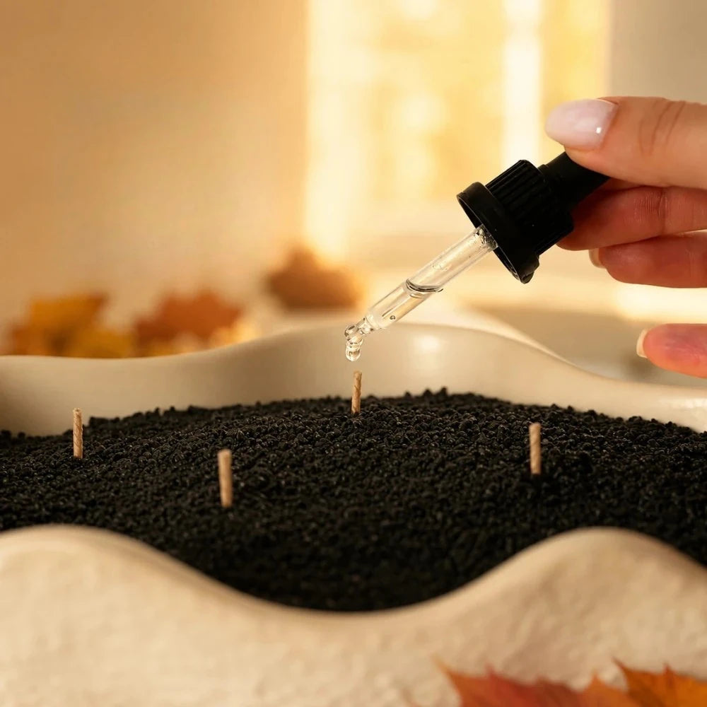 A hand adding pumpkin essential oil with a glass dropper onto black Warmnest candle sand in a wavy cream ceramic dish