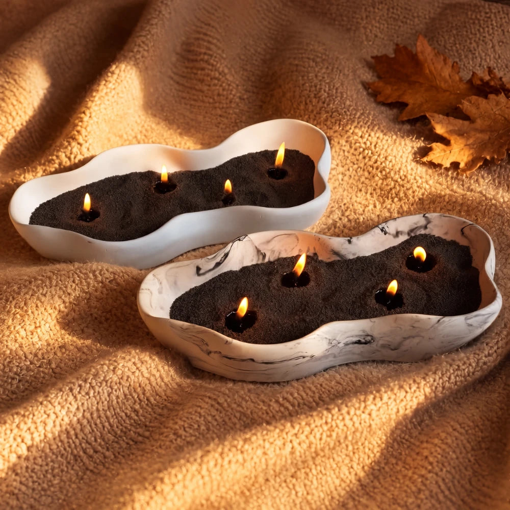 Two wavy ceramic dishes filled with black Warmnest candle sand and lit wicks on a warm caramel wool throw